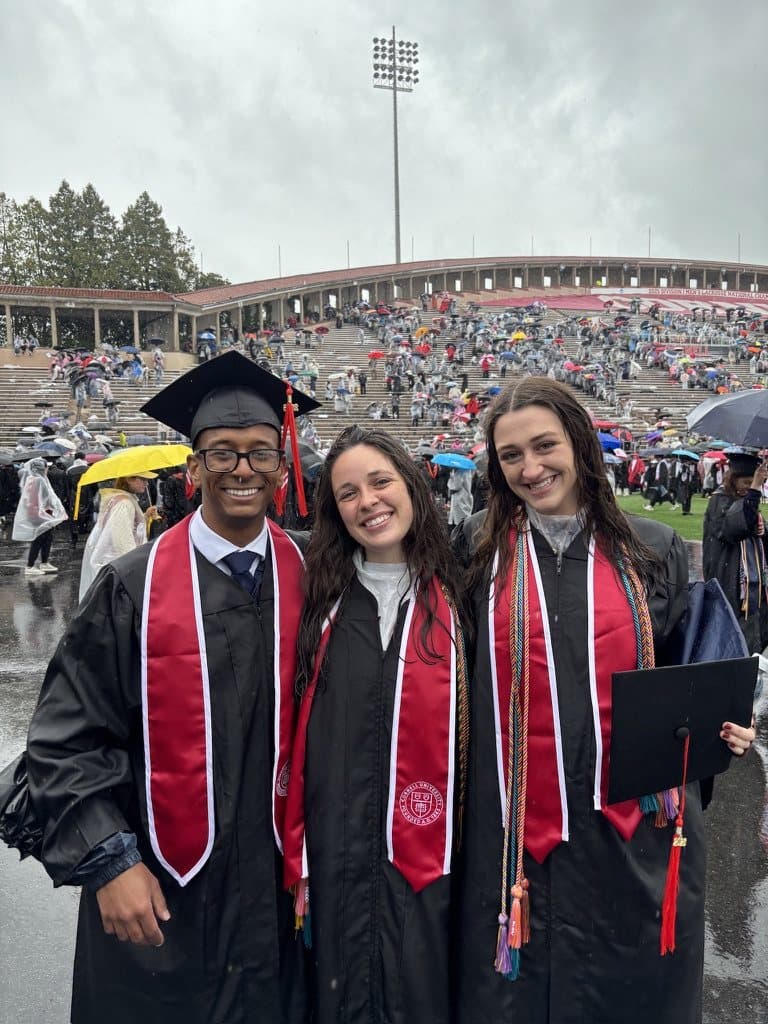 Remy and two friends in caps and gowns at a rainy commencement, with umbrellas filling the stadium